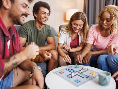 Group of cheerful young friends having fun playing ludo board game while spending time together at home
