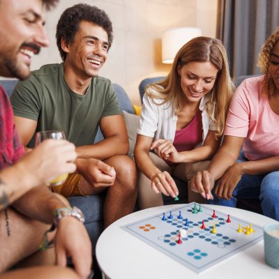 Group of cheerful young friends having fun playing ludo board game while spending time together at home